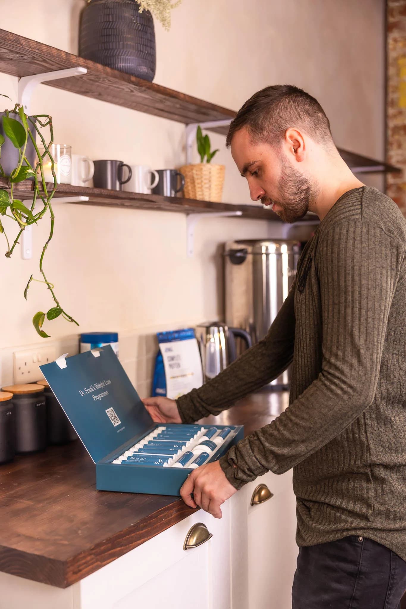 A man in his kitchen in morning light, open shelves of mugs and a trailing plant behind him.