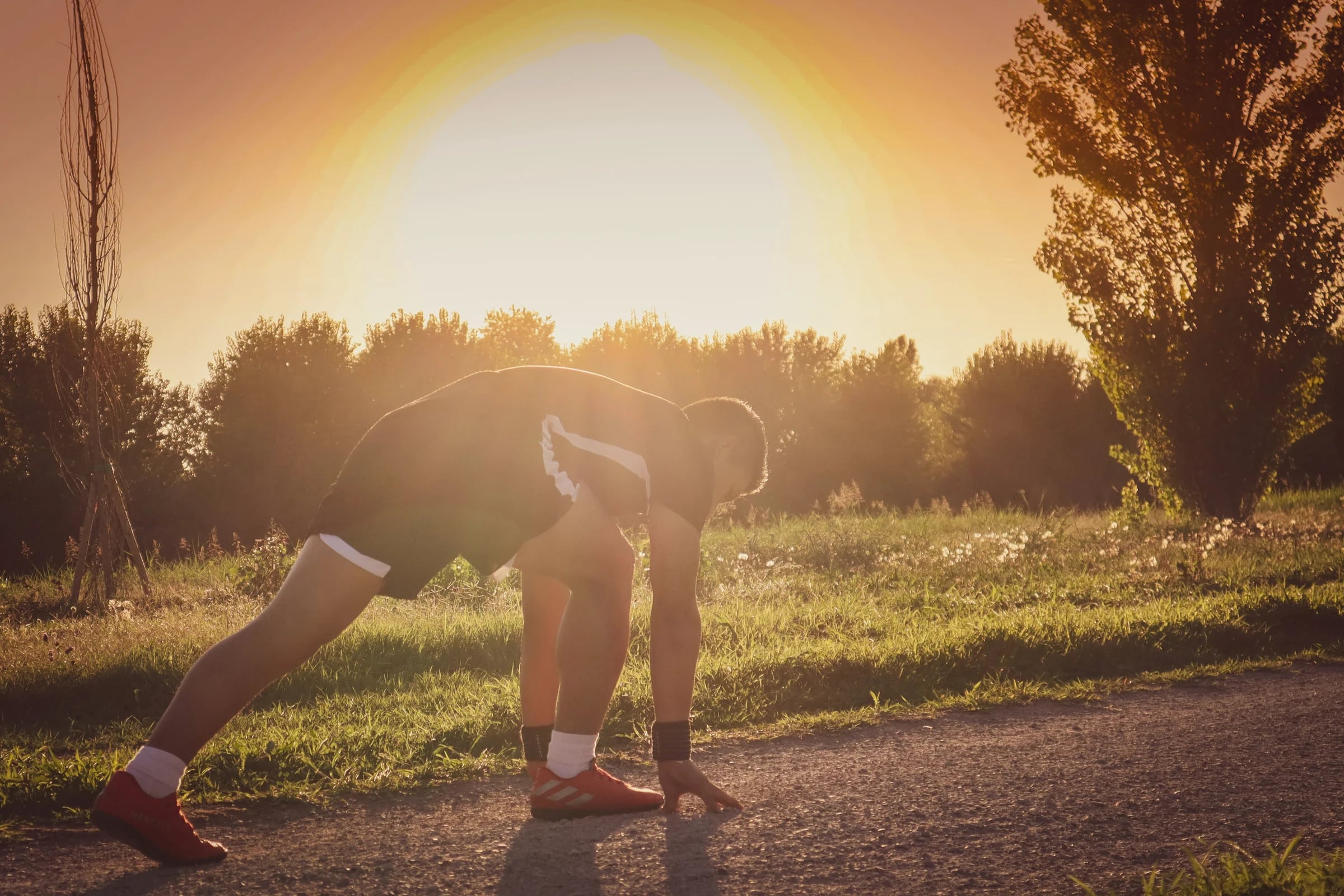A runner stretching on a path as the sun sets behind the trees