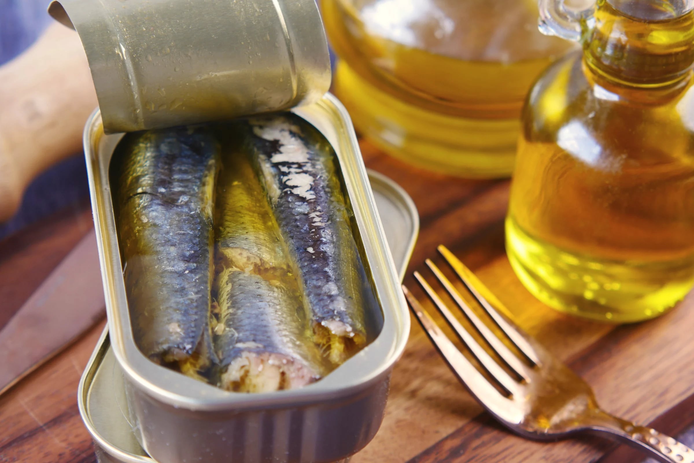An open tin of sardines beside bottles of golden oil and a fork