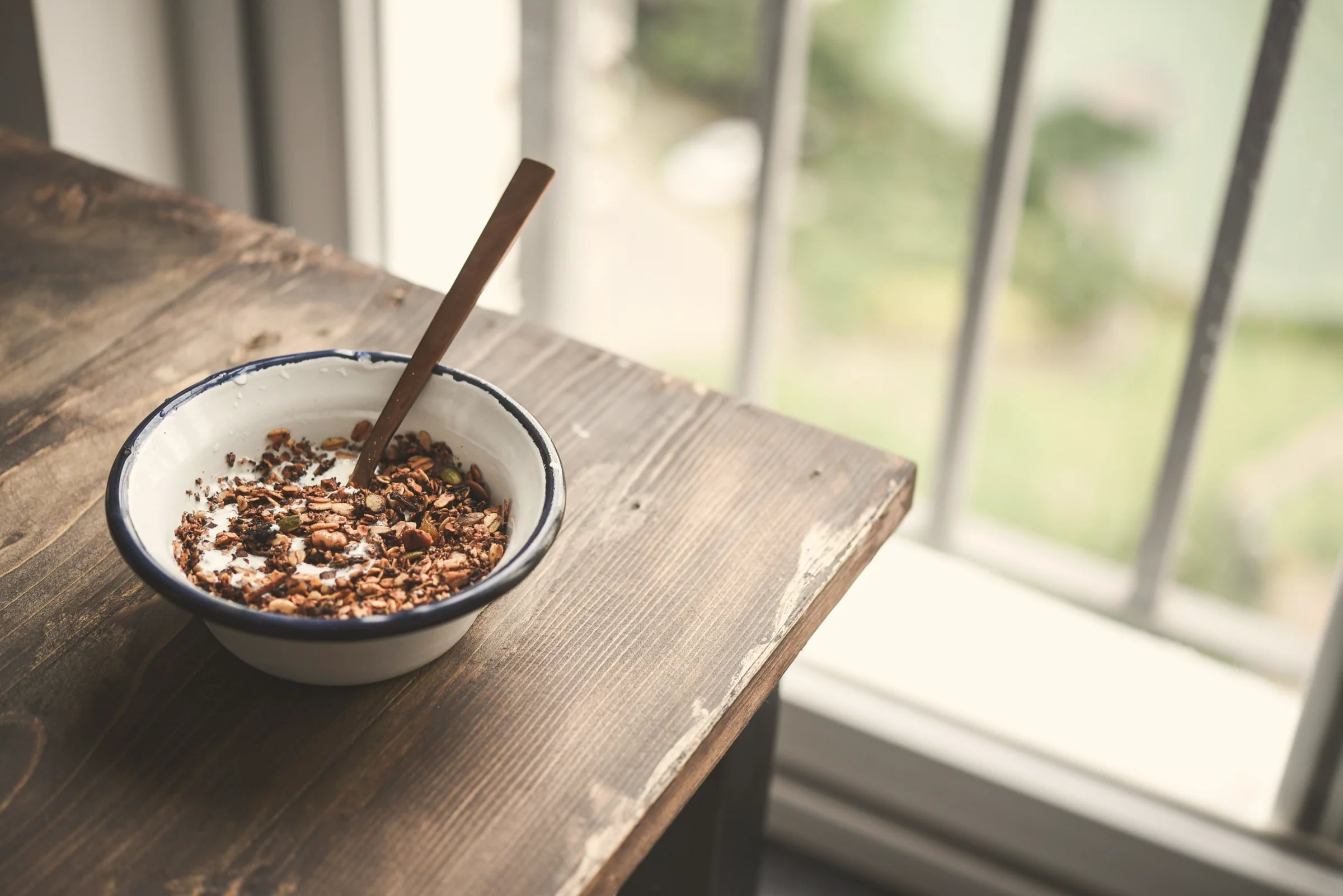A bowl of granola and yoghurt on a wooden table by the window