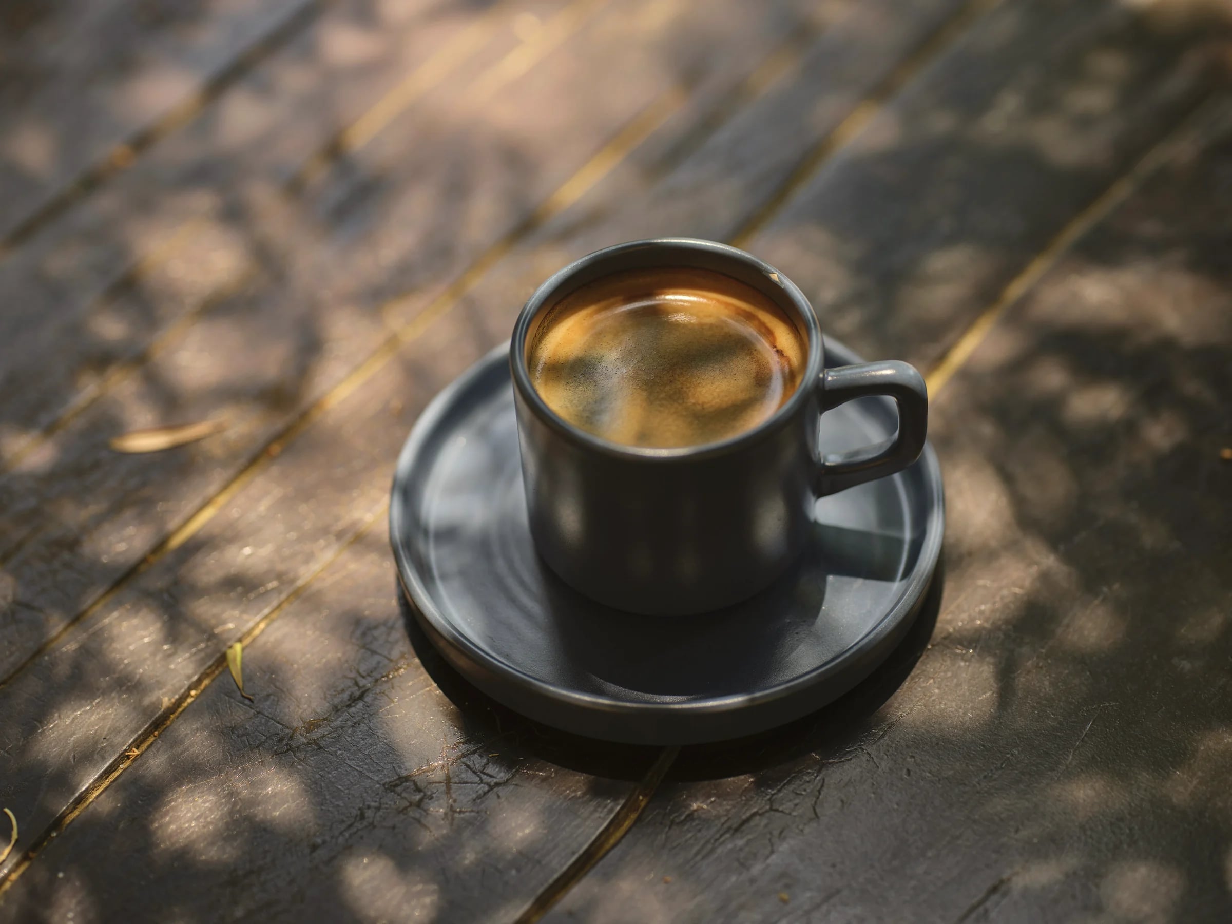 A small cup of black coffee on a saucer in dappled sunlight