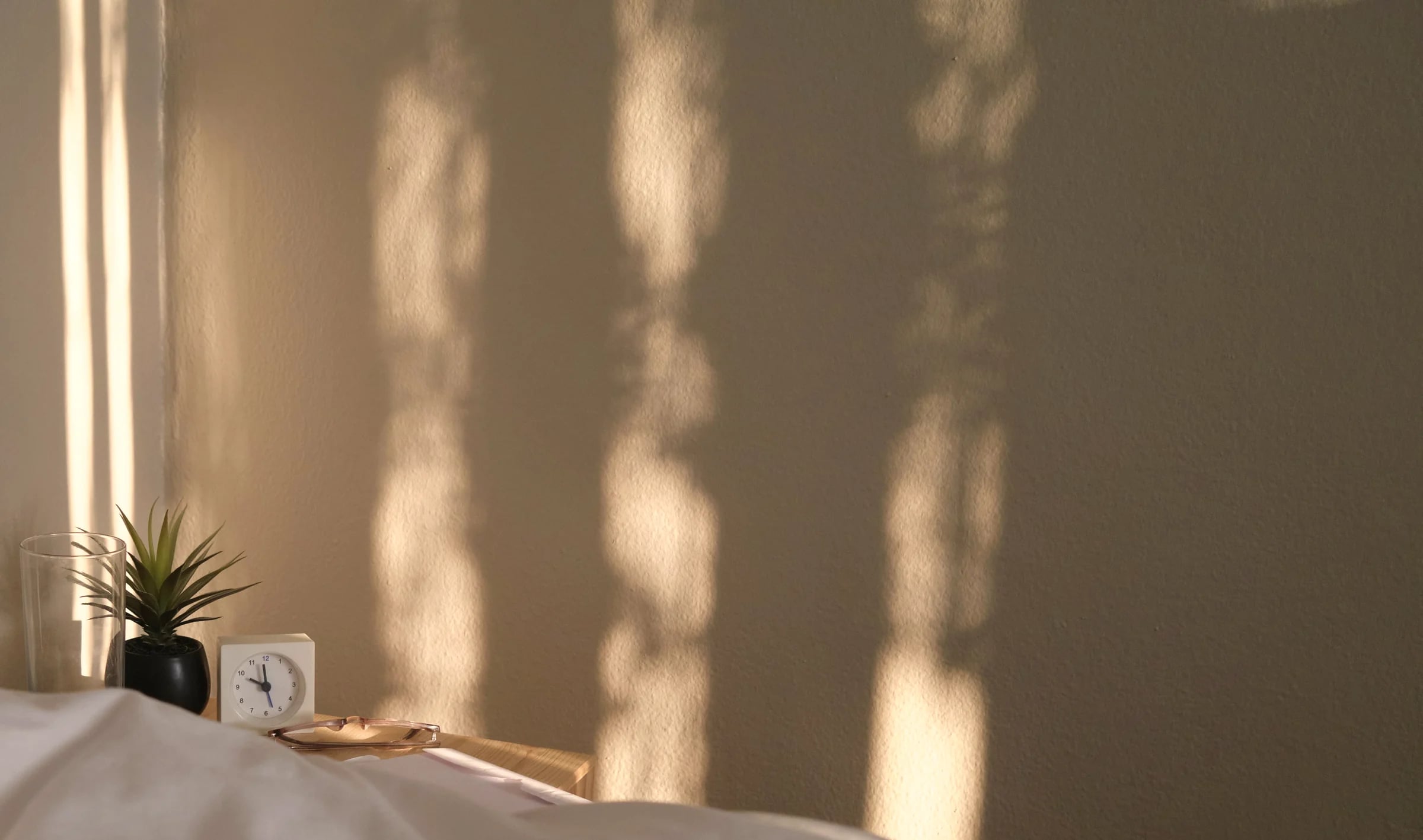 Evening sun casting soft stripes on a bedroom wall above a small clock and a plant